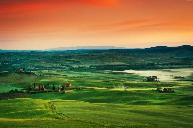 Mist-filled green Tuscan valleys beneath a vivid crimson and orange dawn sky