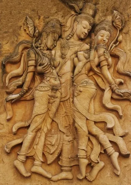 Three crowned celestial dancers in flowing robes, carved in a warm ochre sandstone relief