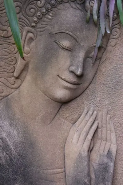 A serene, smiling deity with hands joined in prayer, carved in soft grey stone and framed by palm fronds