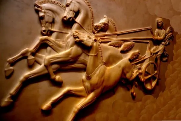Rearing horses pull a charioteer at full gallop, sculpted in glossy copper-brown relief