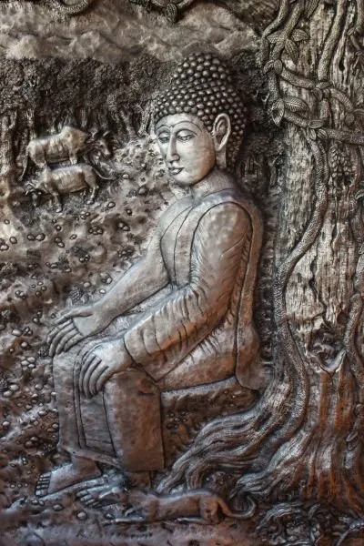 The Buddha sits in quiet reflection beside a great tree trunk, carved in silvery grey stone