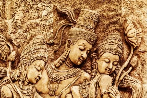 Three crowned figures with serene faces and ornate jewellery, carved in warm golden sandstone with lotus blooms