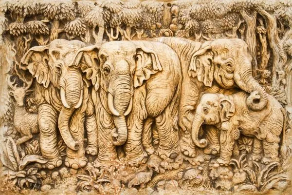 A parade of elephants and calves beneath a canopy of trees, in a pale sandstone relief
