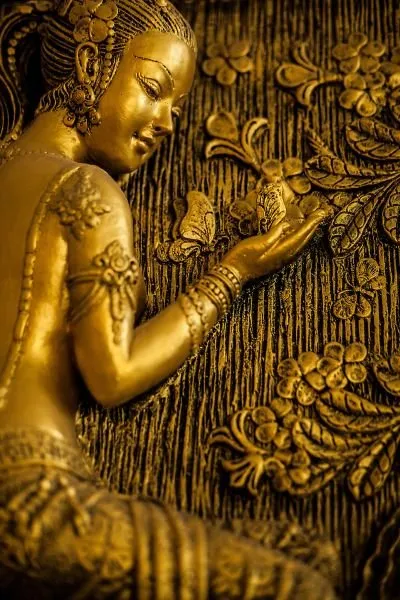 A golden maiden lifts a butterfly to her face beside a tree trunk full of carved blossoms