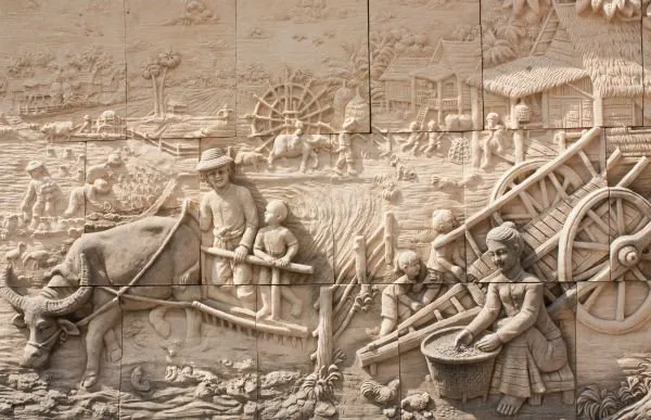Farmers plough with a buffalo and load a cart beside a water wheel, in pale sandstone relief