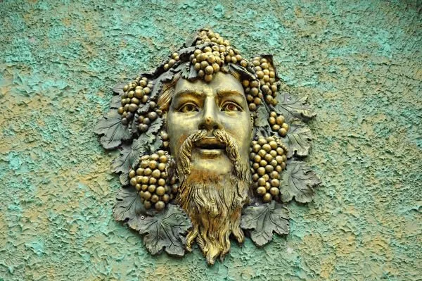 A bearded mask crowned with grapes and vine leaves, in gold and moss green on a turquoise wall