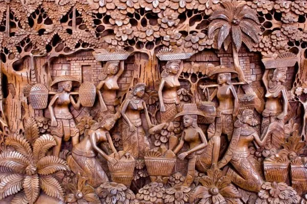 Villagers carry baskets beneath palms and trees in a densely carved, reddish-wood relief