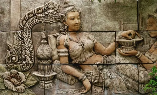 A seated maiden holds out an offering bowl beside a carved serpent and urn on a stone-block wall