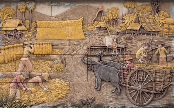 Farmers cut and carry the rice harvest while buffalo pull a cart past thatched huts, in painted relief