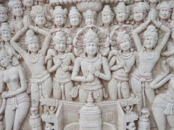 Rows of crowned devotees with joined hands gather around a central shrine, in pale grey stone
