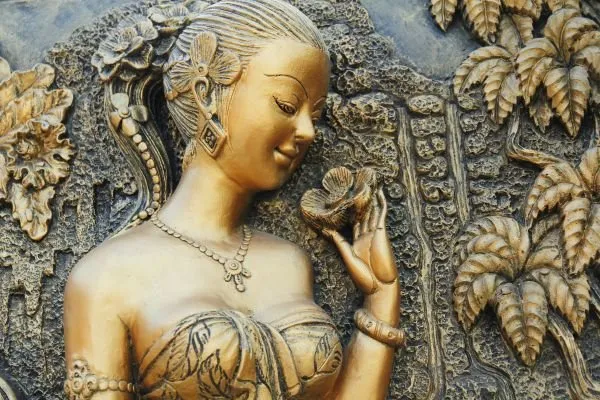 A golden lady with braided hair gazes at a blossom in her hand, beside a carved waterfall garden