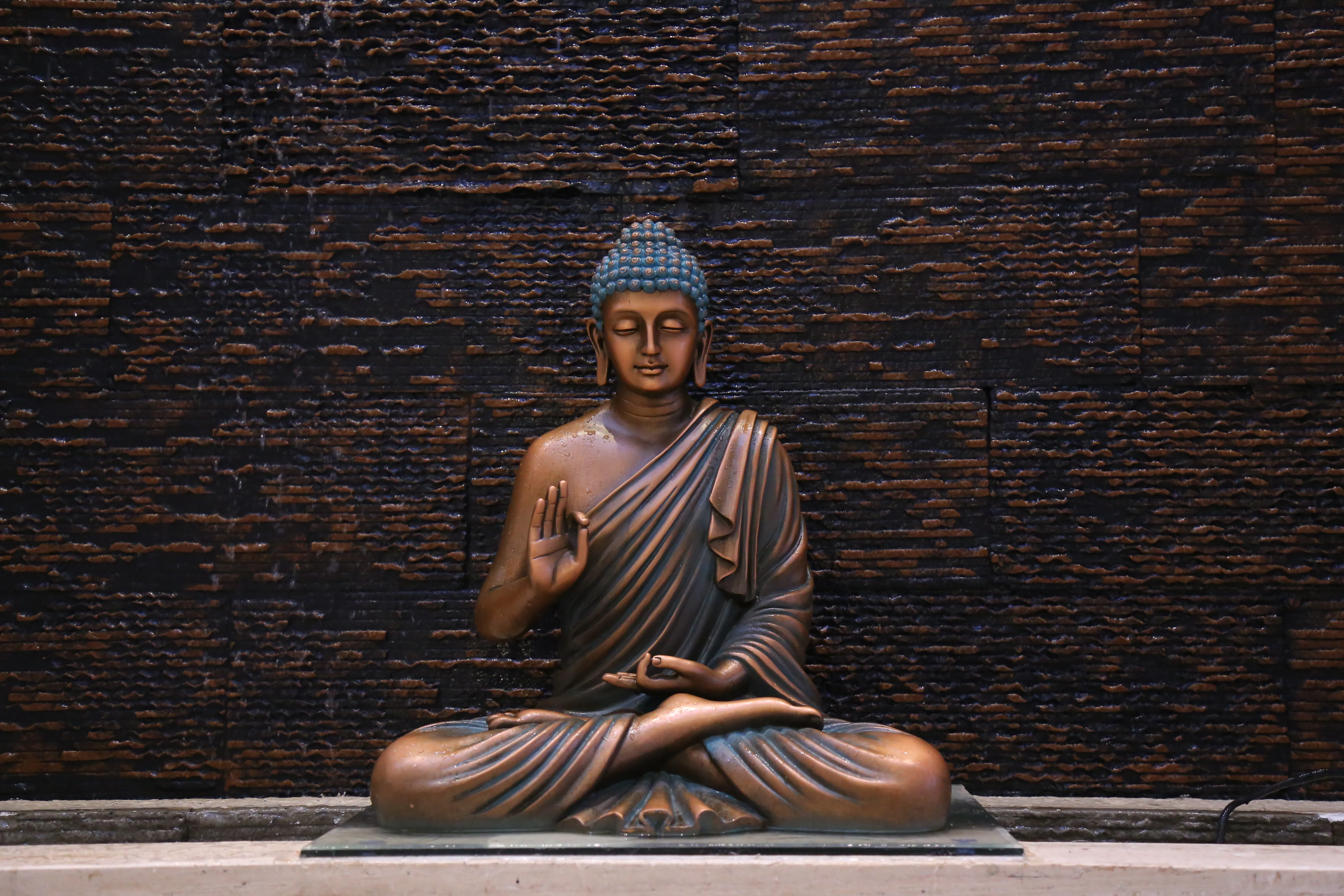 A photograph of a seated bronze Buddha in meditation, one hand raised, before a dark brick wall