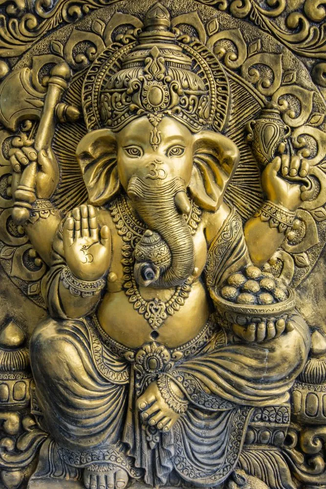An antique-gold embossed-style Ganesha holding a bowl of modak, set within an ornate carved surround