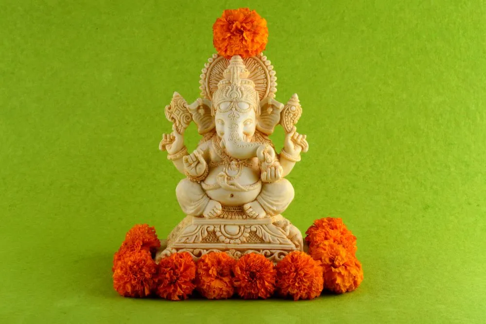 A cream Ganesha idol with a marigold crown and garland, photographed on a bright lime-green background