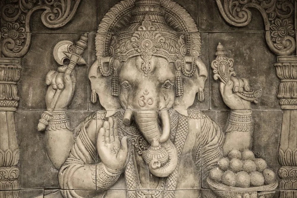 A photograph of a grey stone temple relief showing Ganesha holding a bowl of modak