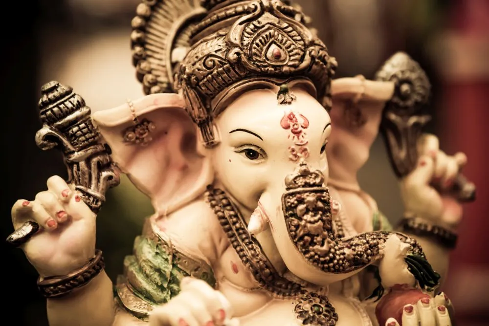 A close-up photograph of a painted festival Ganesha idol with an ornate crown and soft pink tones