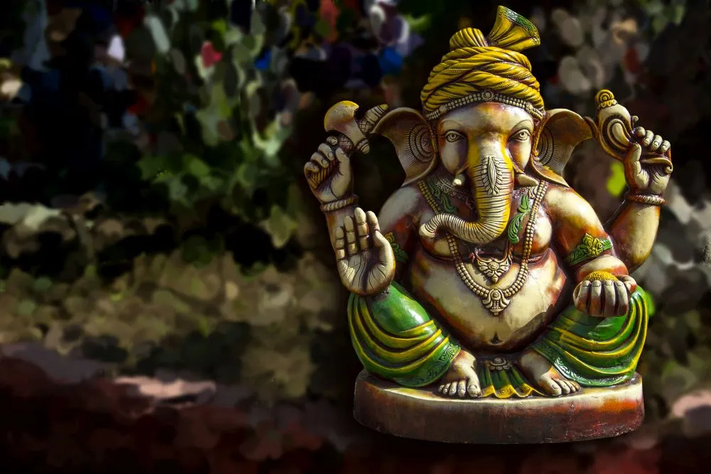 A photograph of a painted Ganesha idol in a yellow turban, set outdoors against soft green foliage