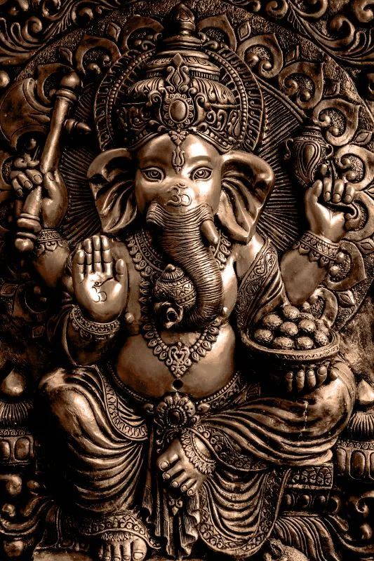 A seated Ganesha in warm antique-bronze relief, holding a modak bowl within ornate carved scrollwork