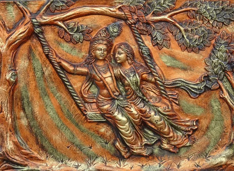 Radha and Krishna on a swing beneath a spreading tree, rendered as a burnished copper relief